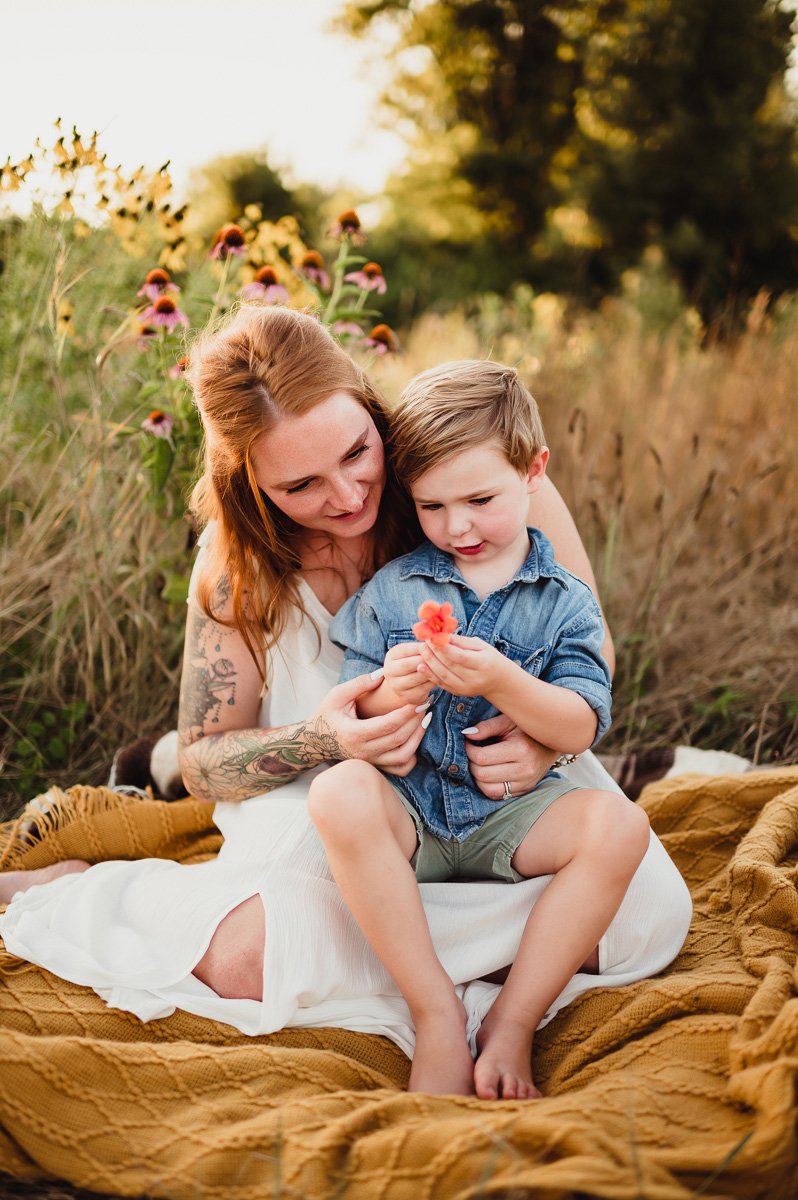 mother and toddler son sitting together admiring a flower in a midwest summer field