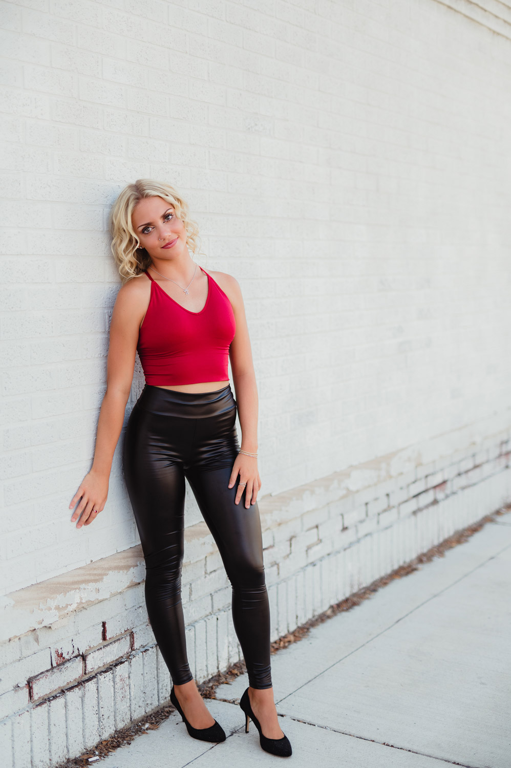 senior girl in red tank top and black leather plants standing in front of white brick wall
