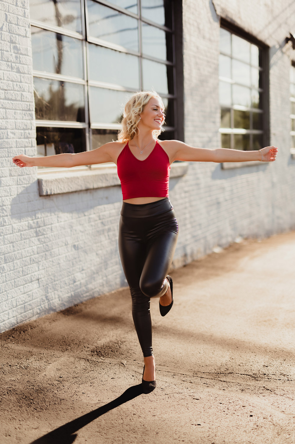 senior girl in red tank top and black leather plants, arms outstretched and one leg up, standing in front of white brick wall