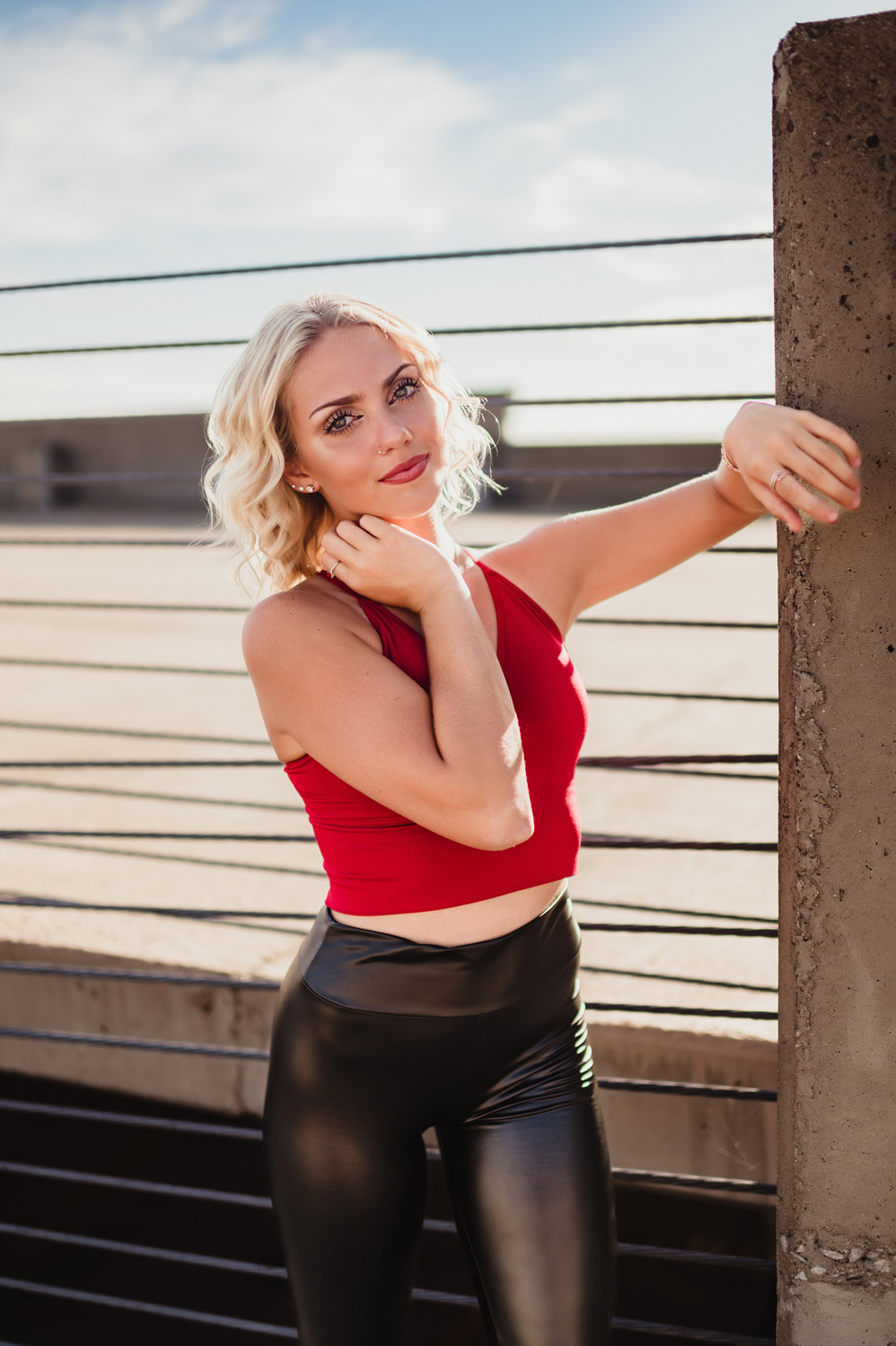 senior girl in red tank top and black leather plants posing on the rail of a parking deck in Lafayette, IN