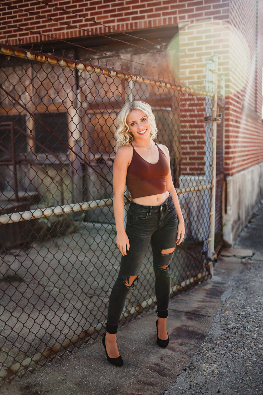 senior girl in standing pose in front of chain link fence outside of an urban warehouse