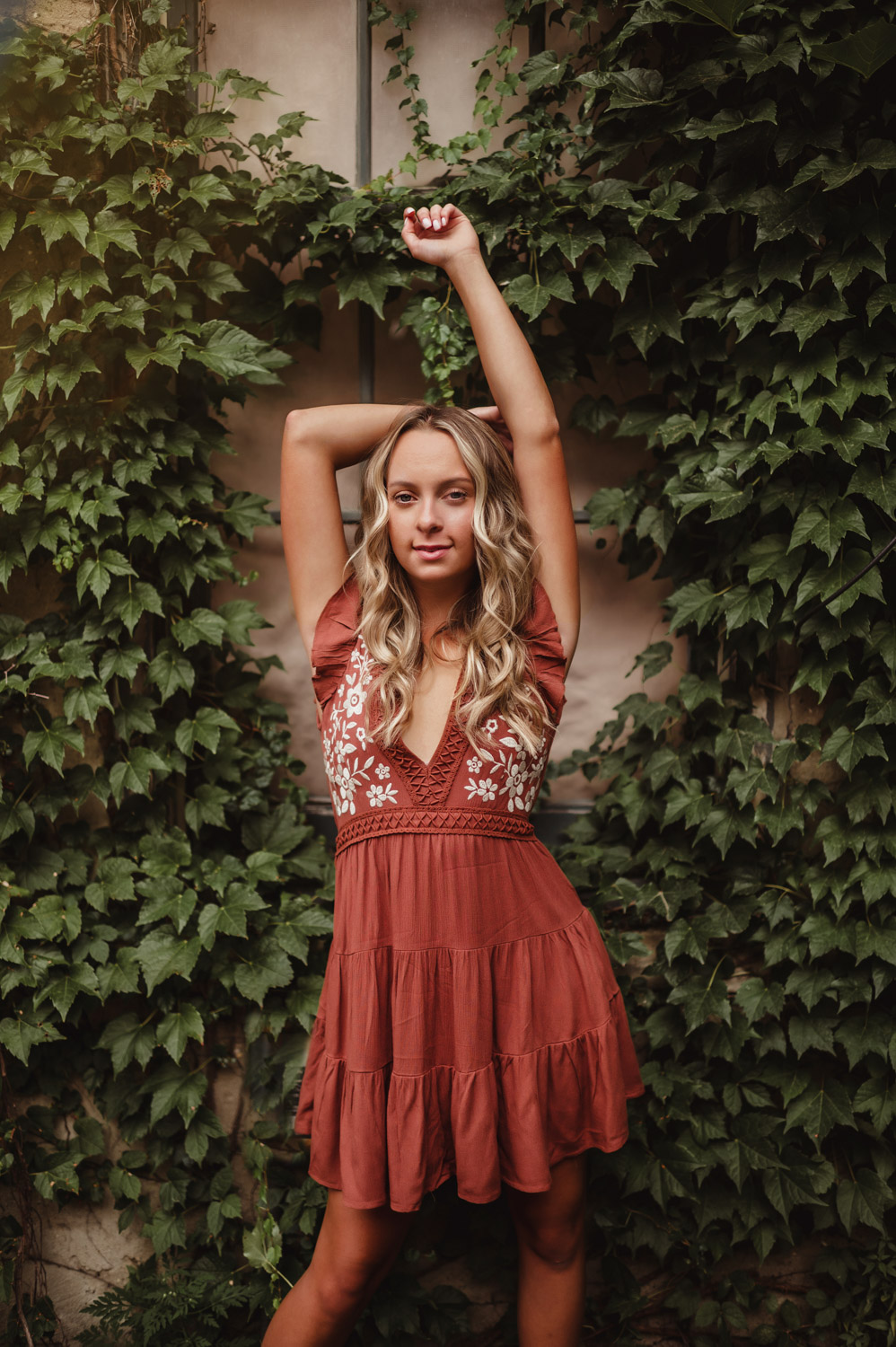 How to Get Creamy Skin Tones in Photos-how to get creamy skin tones in photos Young woman with long wavy blonde hair poses in front of a lush green ivy wall, wearing a rust-colored dress with white floral embroidery; soft, natural lighting enhances her creamy skin tones and glowy complexion.