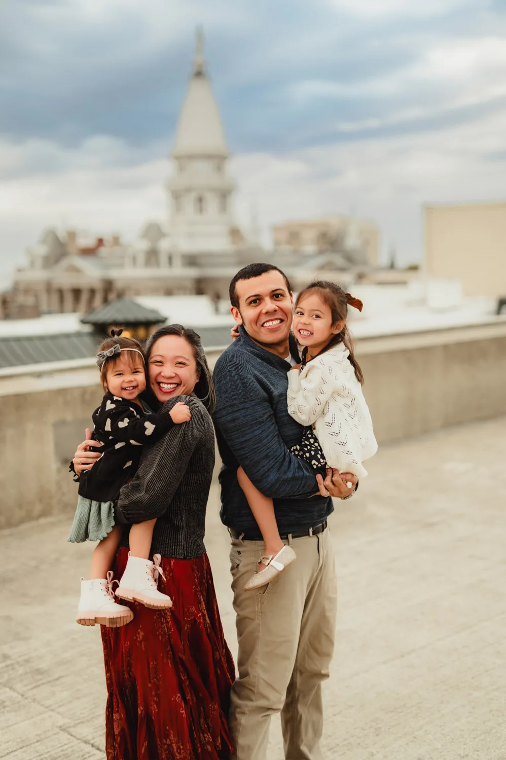 parents posing with two young daughters  on top of urban parking garage with county courthouse in background