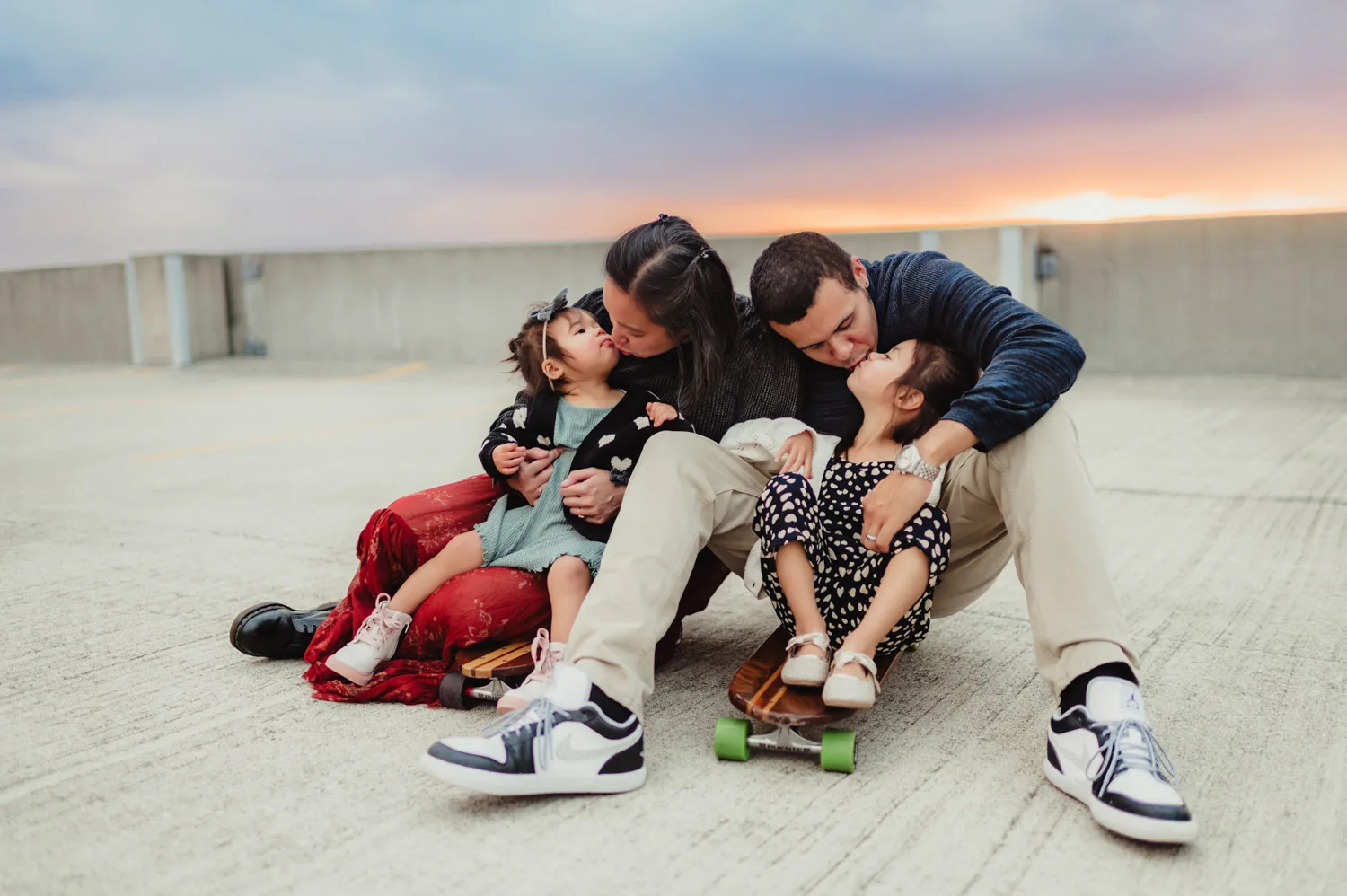 mom and dad sitting on two skateboards kissing their two young daughters
