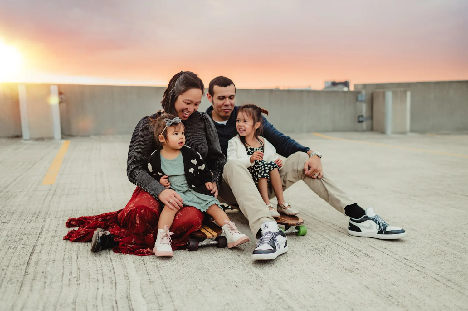 mom and dad sitting on two skateboards with two young daughters on top of urban parking garage at sunset
