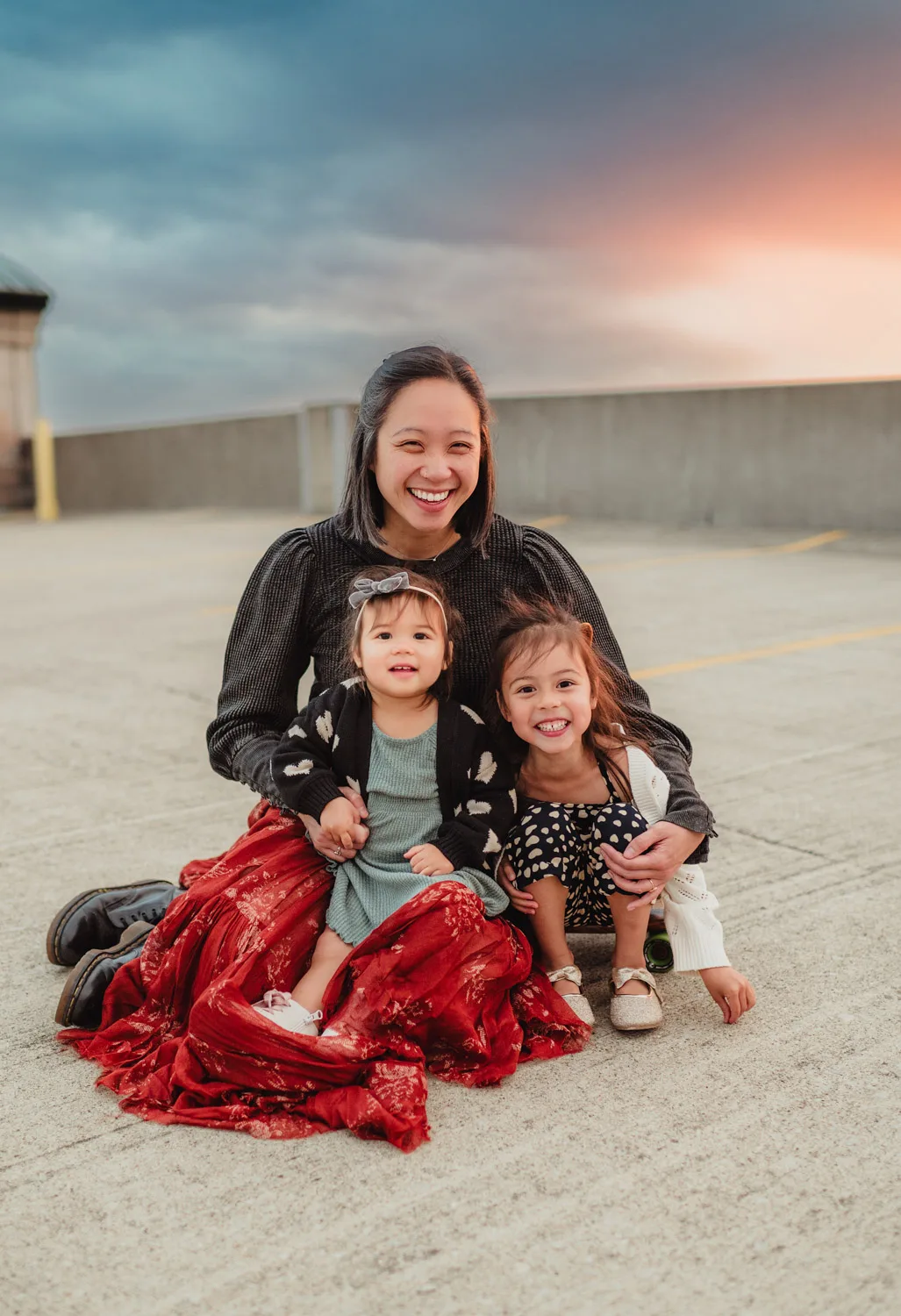 mom sitting and holding two young daughters in parking lot at sunset