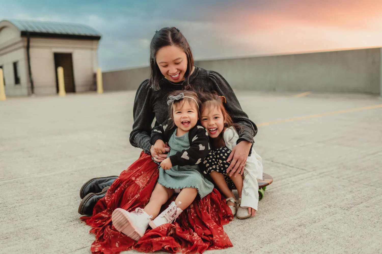 mom sitting and holding two young daughters on top of parking garage at sunset