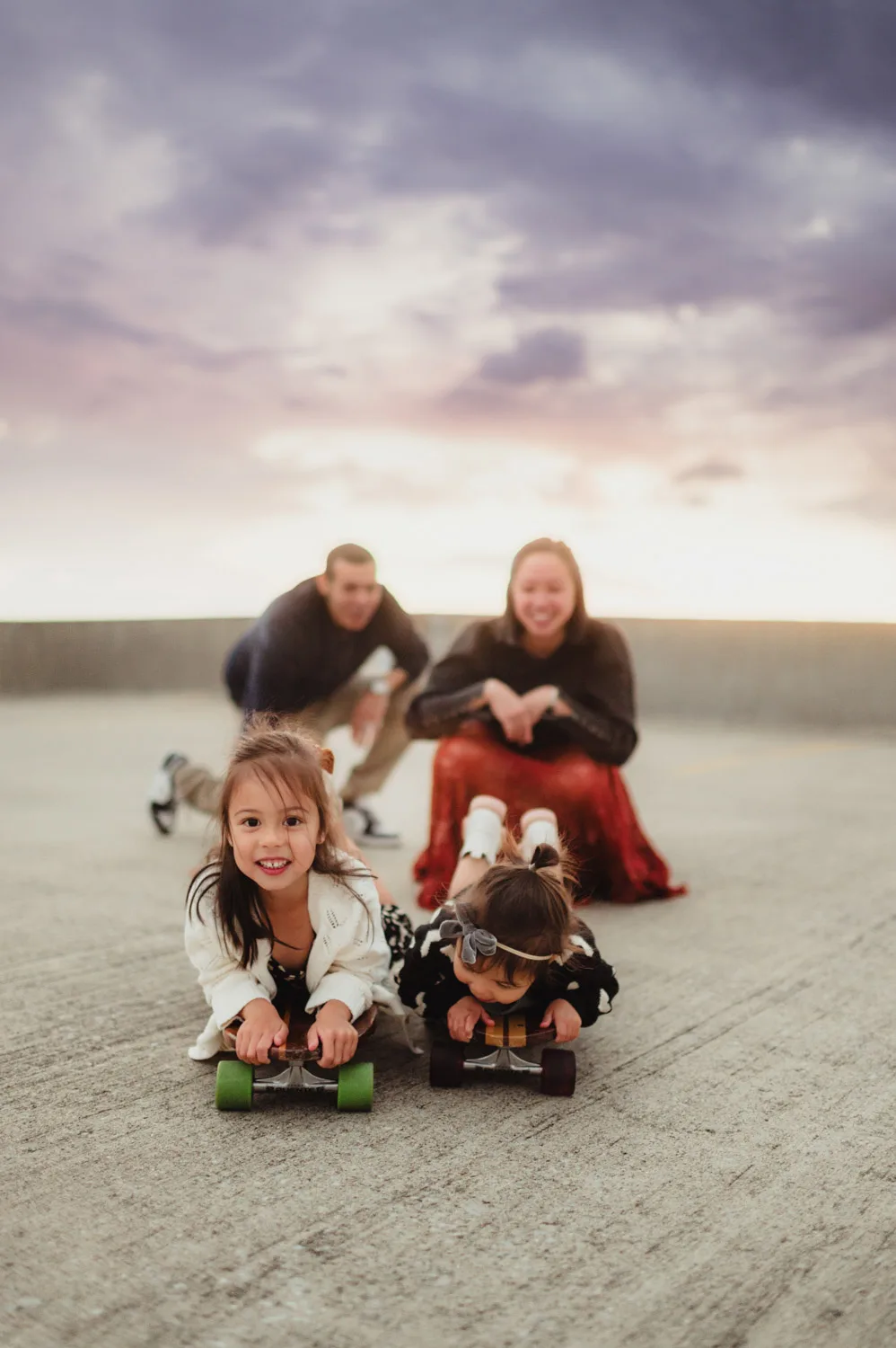 two young girls laying face first on skateboards with parents looking on in background at sunset