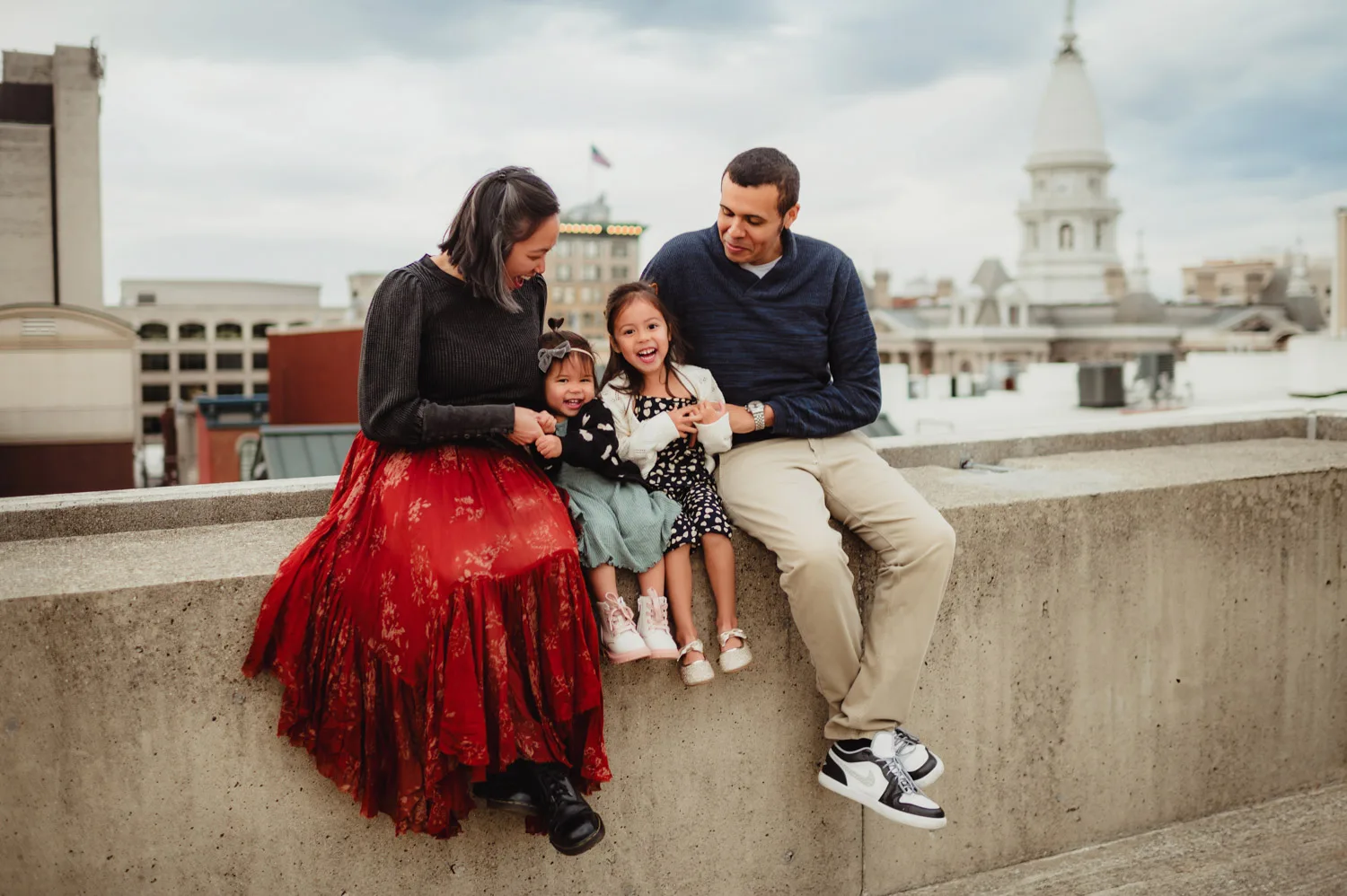 parents sitting and laughing on wall of urban parking garage with two young daughters with courthouse in background