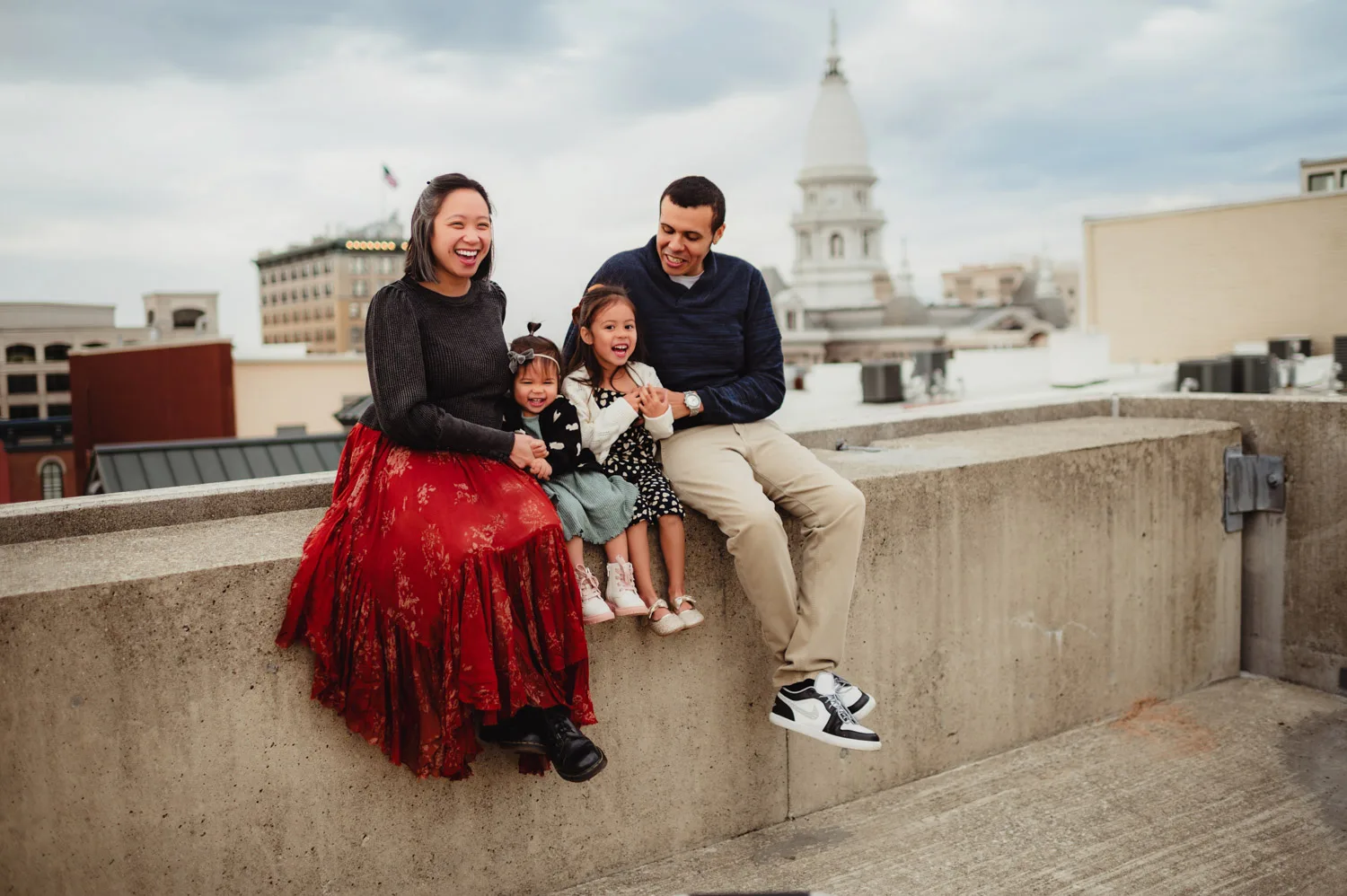 parents sitting on wall of urban parking garage with two young daughters with courthouse in background