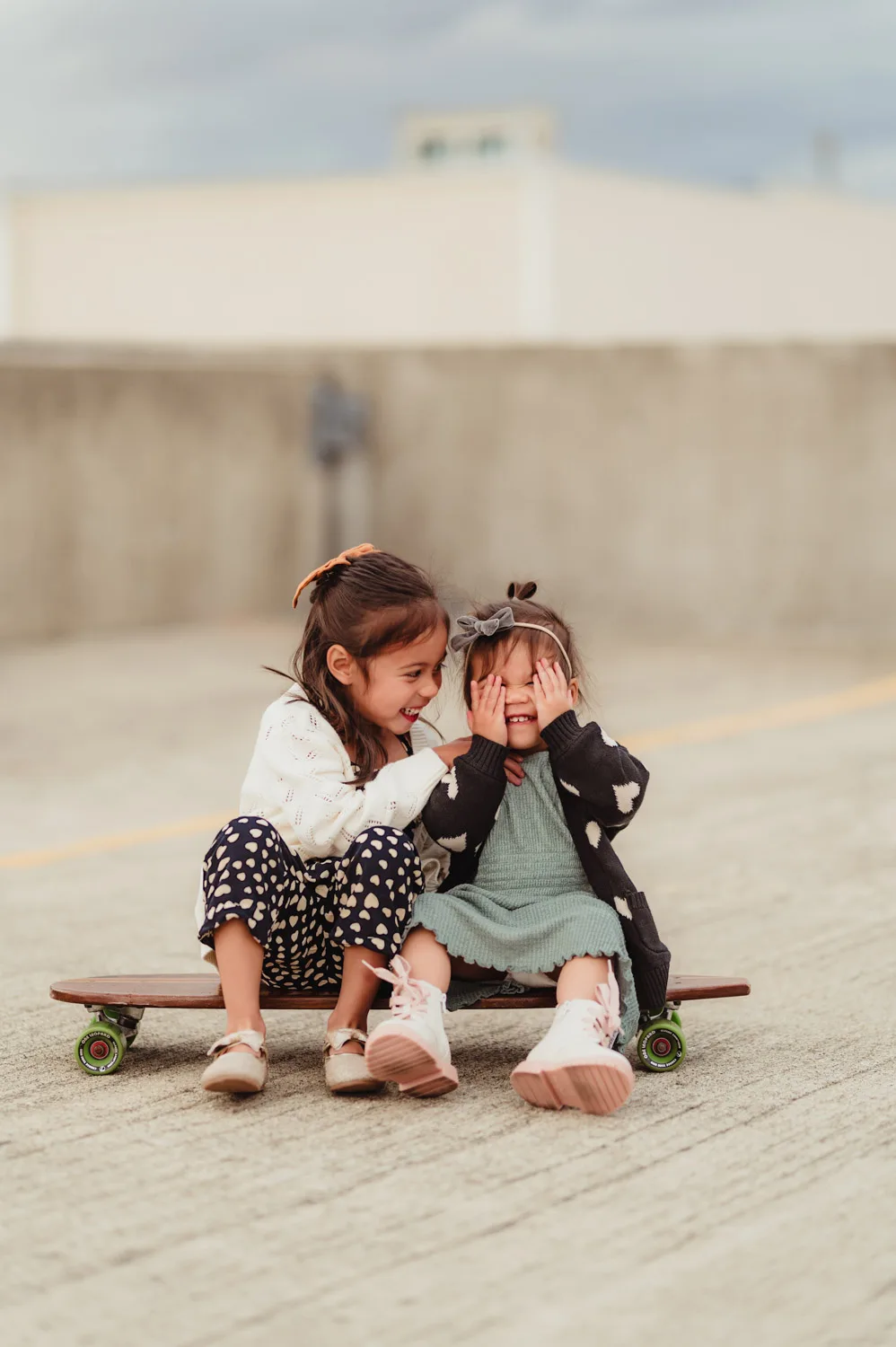 two young sisters playing peek-a-boo on skateboard together on top of urban parking garage