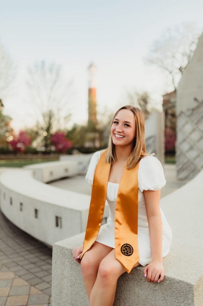 College Grad Photos: 5 Must-Know Tips for an Unforgettable Photo Session-college grad photos College graduate in a white dress and gold stole sitting on a modern concrete monument, smiling with spring trees and campus in the background.