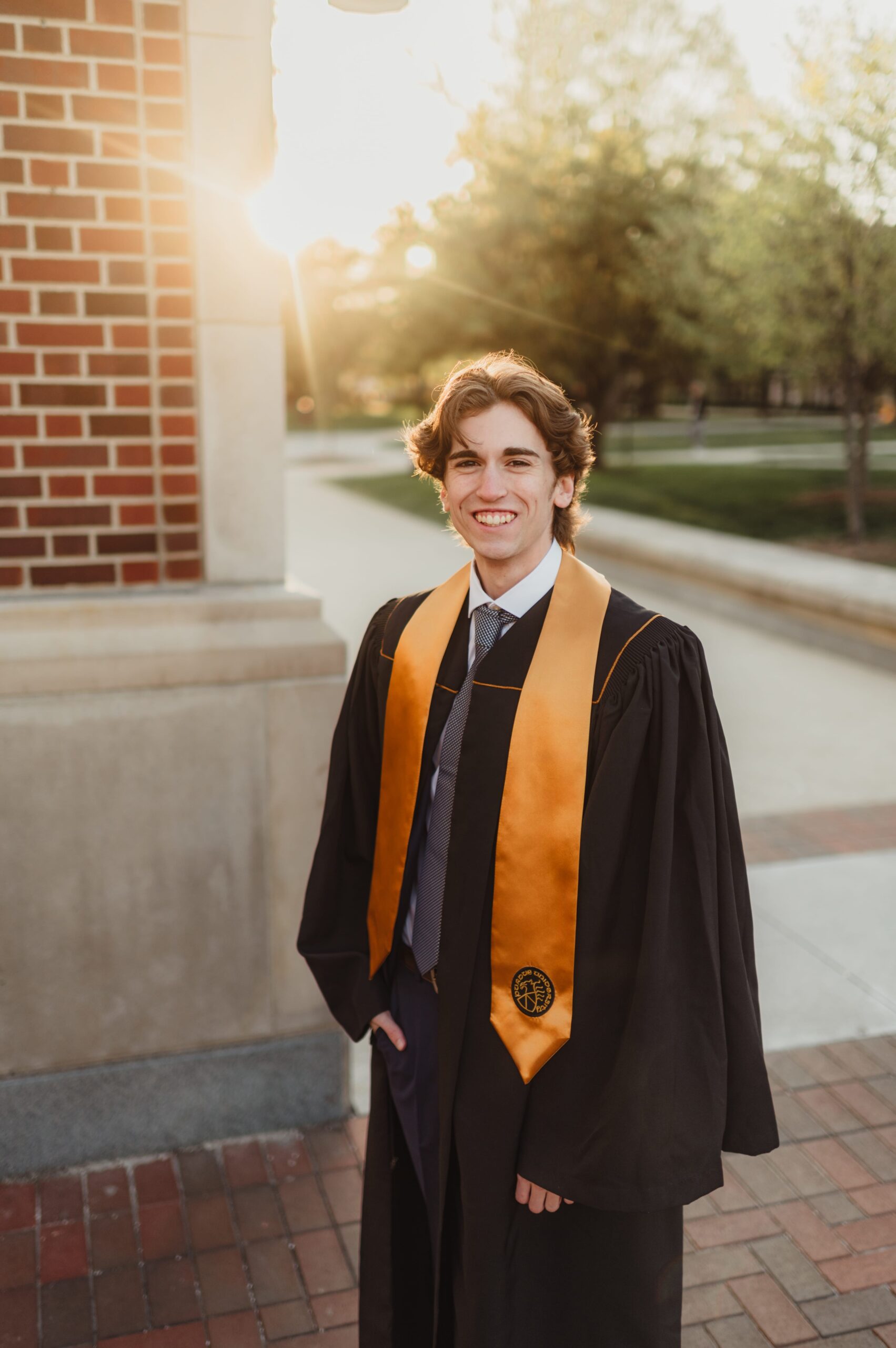 Male graduate in gown at golden hour Male college graduate in a black cap and gown with a gold stole, smiling at golden hour near a brick campus building.