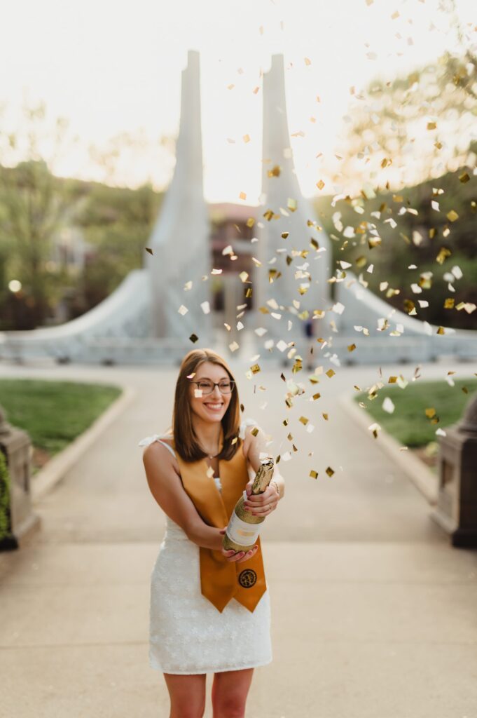 College Grad Photos: 5 Must-Know Tips for an Unforgettable Photo Session College graduate wearing a white dress and gold stole pops champagne with gold confetti flying in front of campus fountain at sunset.