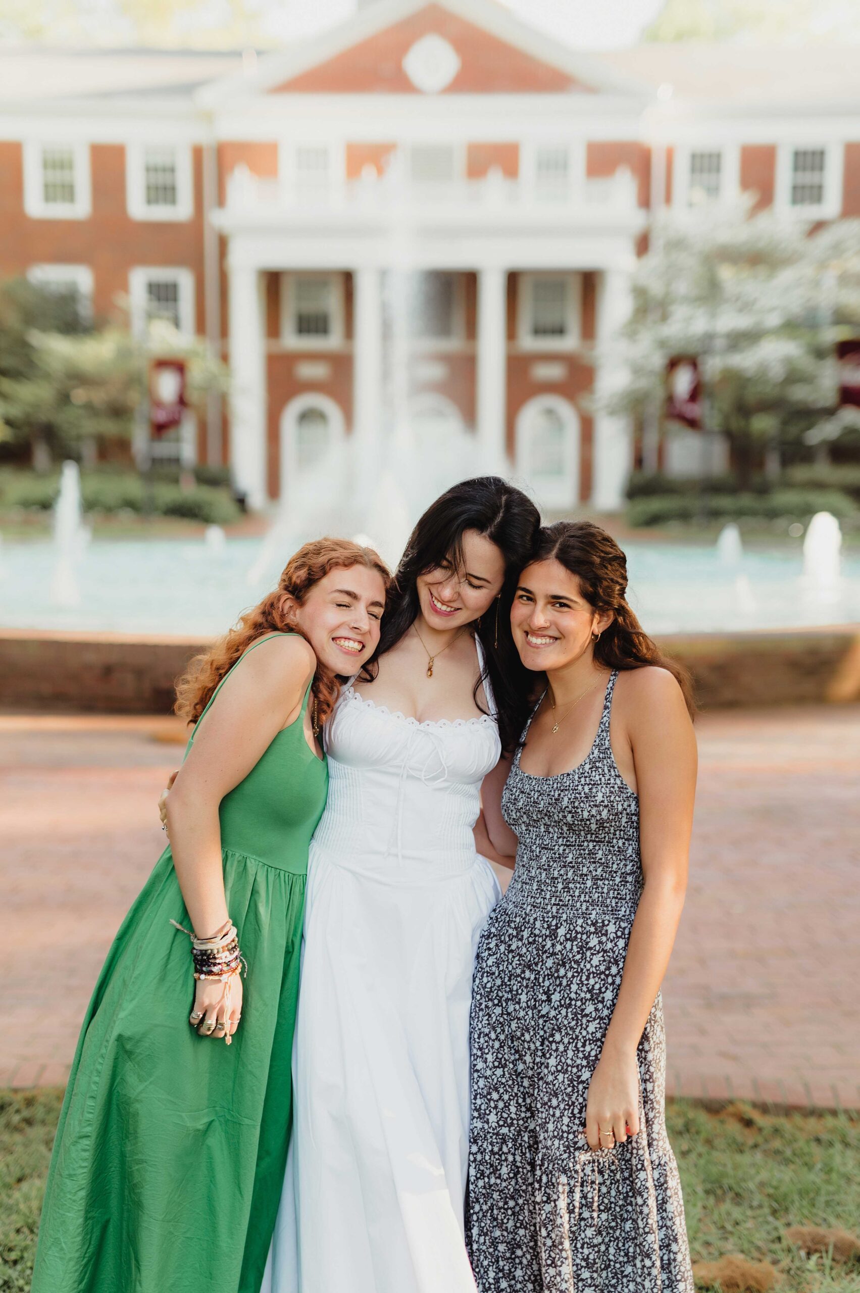 College Grad Photos: 5 Must-Know Tips for an Unforgettable Photo Session Elon University graduate in a white dress smiling and hugging two close friends in front of a campus fountain and brick building.”