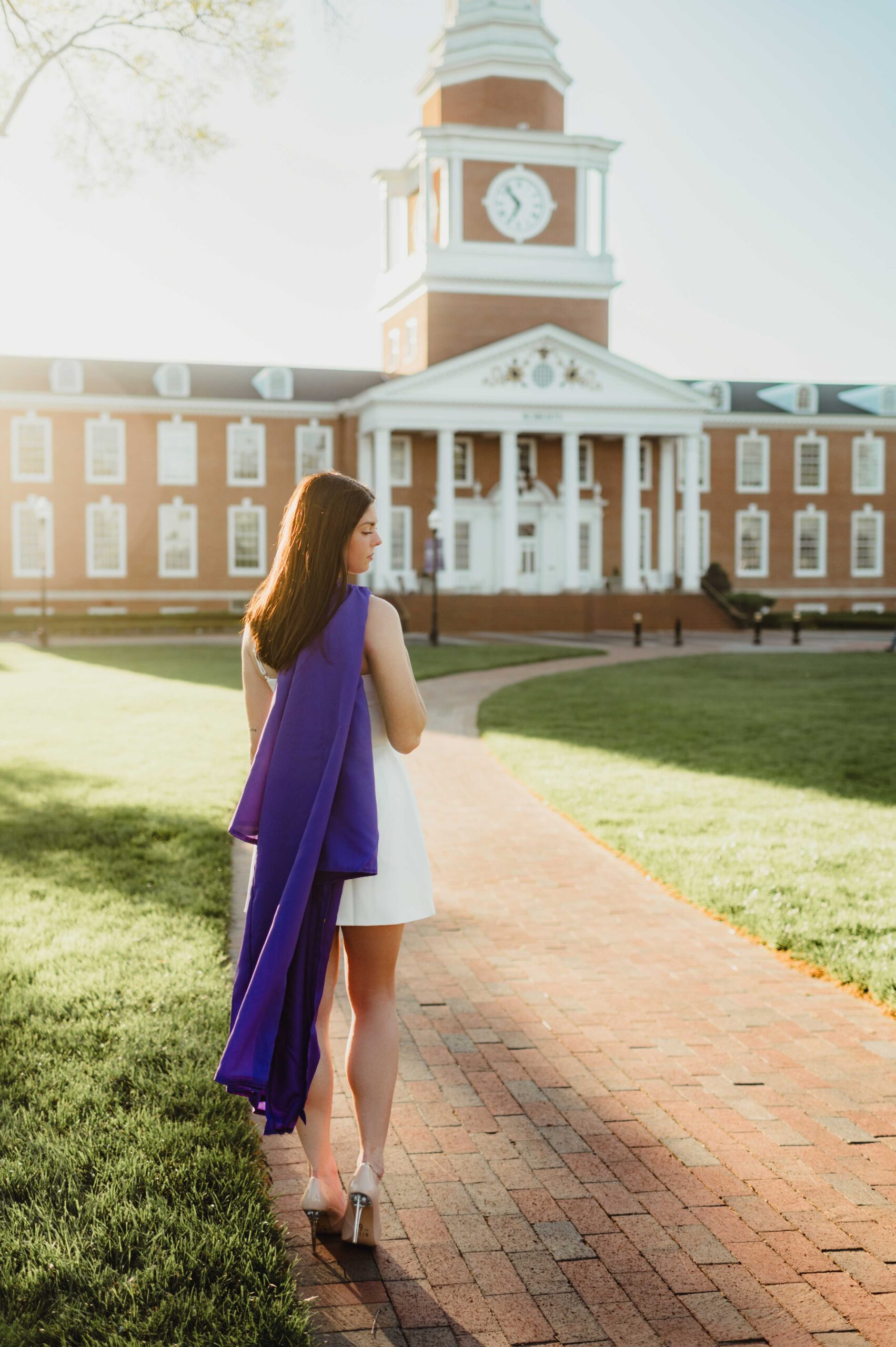 College Grad Photos: 5 Must-Know Tips for an Unforgettable Photo Session College graduate in a white dress with a purple graduation gown draped over her shoulder, standing on a brick path at High Point University during golden hour.