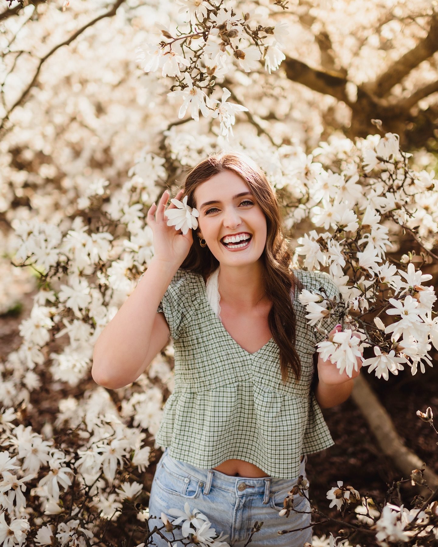 Capture the Real You: A Senior Photo Session Experience in Greensboro-senior photo session in Greensboro Joyful senior portrait in Greensboro NC featuring a high school senior girl in a green gingham top surrounded by blooming white magnolias.