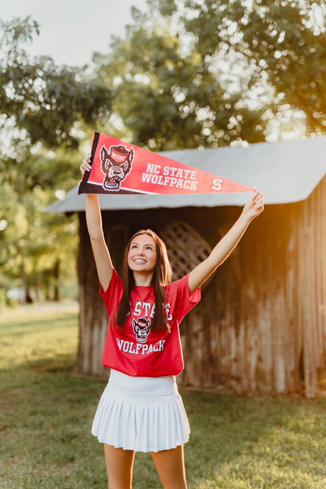 🌼 A Wildflower Field at Golden Hour: Genna’s Senior Session | Greensboro Senior Photographer-Greensboro Senior Photographer High school senior proudly holding NC State Wolfpack pennant in red t-shirt and white skirt, photographed at golden hour.