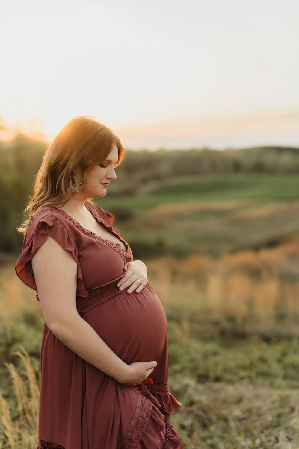 Expecting Baby #2: Golden Hour Maternity Photos in Greensboro-Greensboro maternity photos Pregnant woman in rust-colored dress cradling her belly at sunset during a maternity photo session near Greensboro, NC