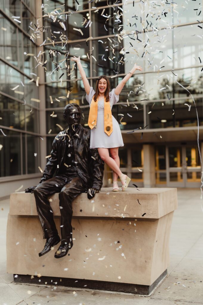 Graduate with confetti standing on statue College graduate standing next to a statue, throwing her hands up joyfully as black, gold, and white confetti rains down in front of a glass building on campus.