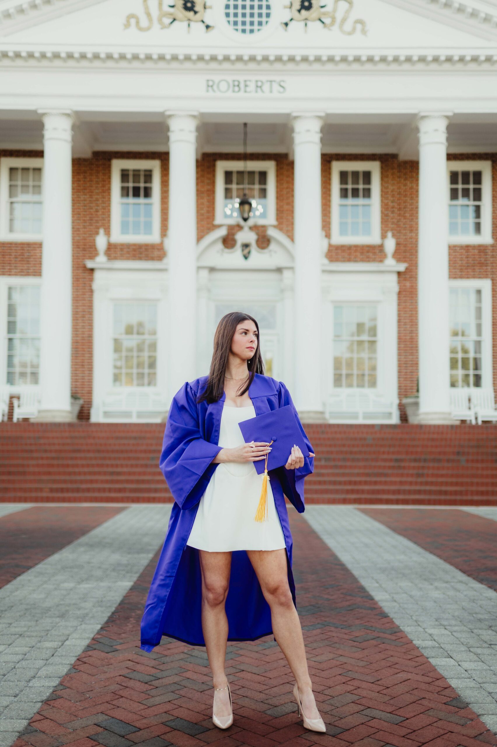 Laura's Graduation Photos at High Point University: Iconic Campus Spots + Champagne Fun-graduation photos at high point university High Point University graduate in cap and gown holding her cap in front of Roberts Hall during senior photo session.