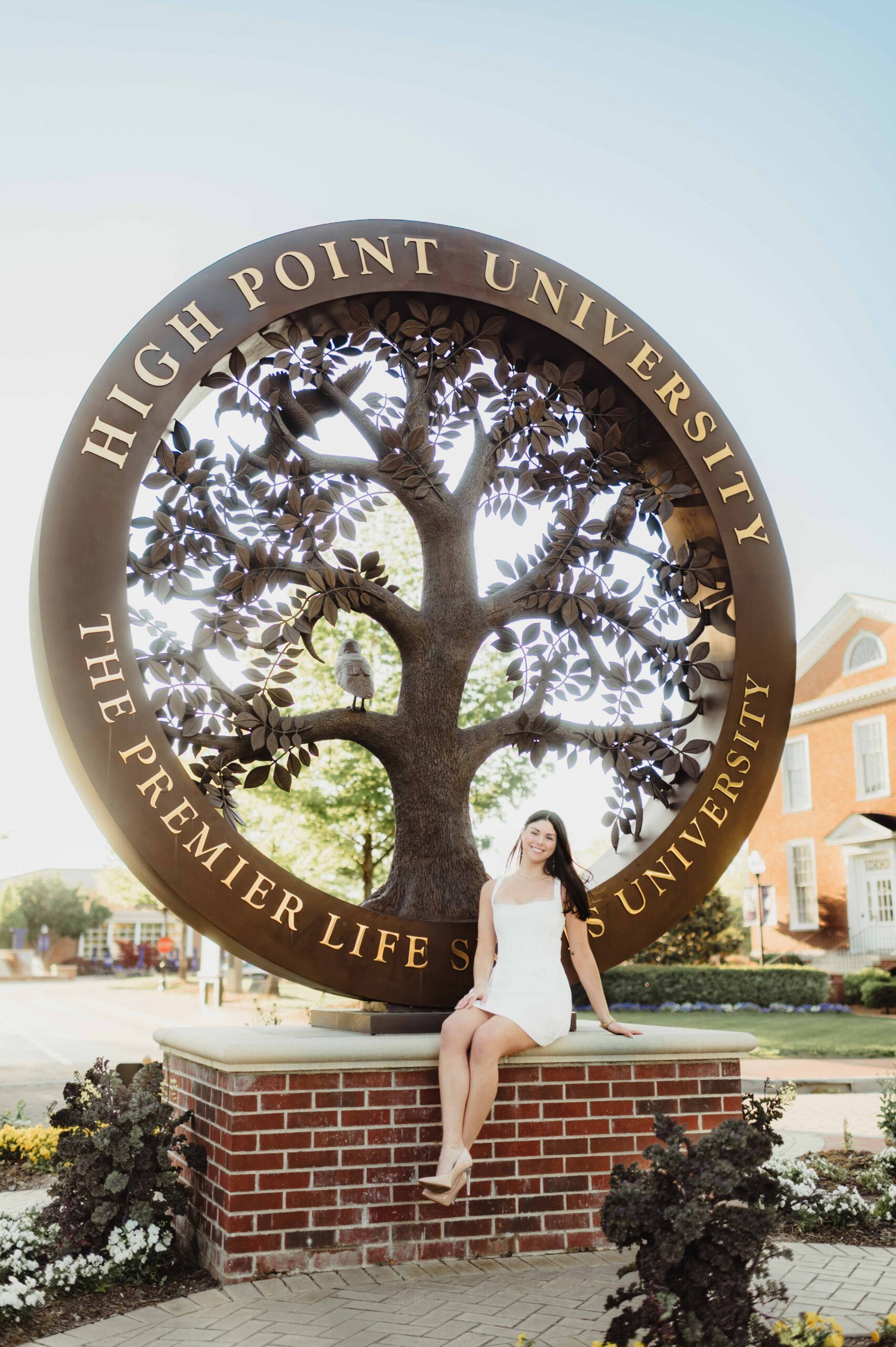 Laura's Graduation Photos at High Point University: Iconic Campus Spots + Champagne Fun-graduation photos at high point university Graduate sitting on the base of the Tree of Life sculpture at High Point University wearing a white dress and heels.
