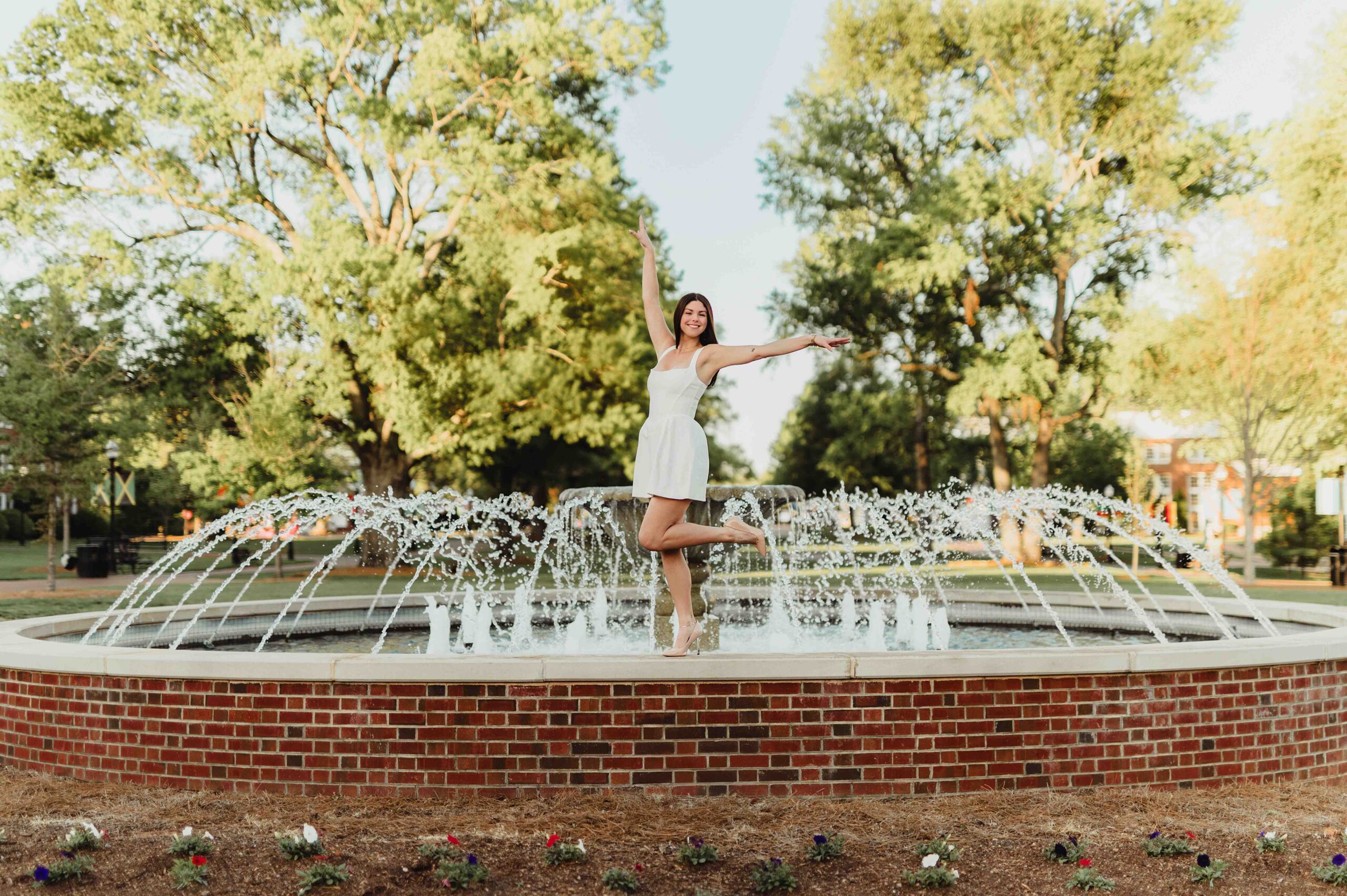 Laura's Graduation Photos at High Point University: Iconic Campus Spots + Champagne Fun-graduation photos at high point university High Point University senior jumping joyfully in front of the Henley Fountain in a white graduation dress.