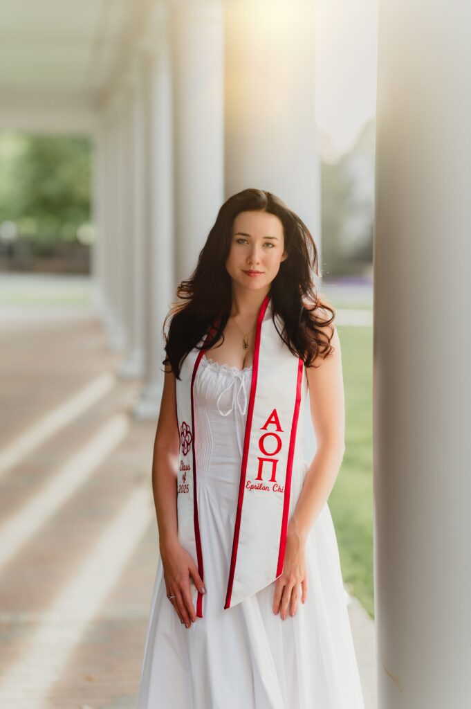 Graduate in sorority stole with white dress College graduate wearing a white dress and red-and-white sorority stole, standing between white columns at Elon University in soft golden light.”
