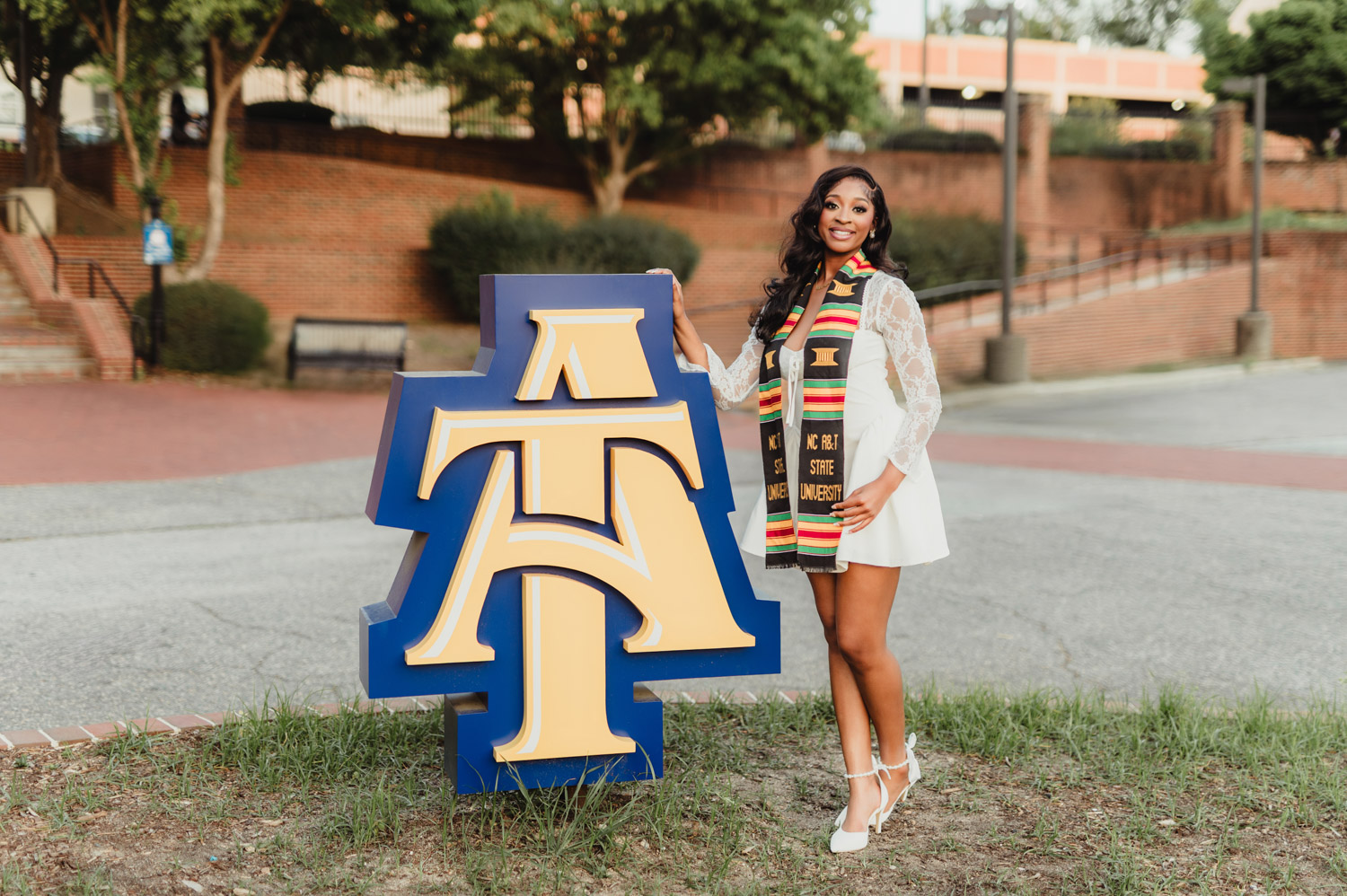 NC A&T Graduation Photos - Victoria’s Golden Hour Session-NC A&T graduation photos NC A&T graduation photo of a young woman smiling beside a large blue and gold A&T sign, wearing a kente stole and white dress.