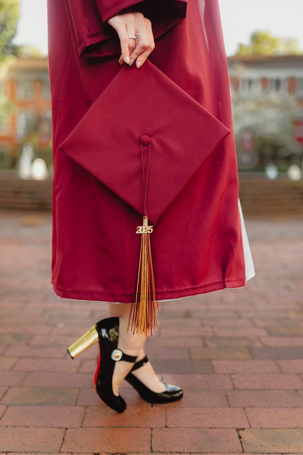Graduation Photos at Elon University: Celebrating Amelia's Big Moment-Graduation Photos at Elon University Close-up of an Elon University graduate holding a maroon cap with a gold 2025 tassel, wearing a gown and black heels with gold block heels on campus brick pathways.
