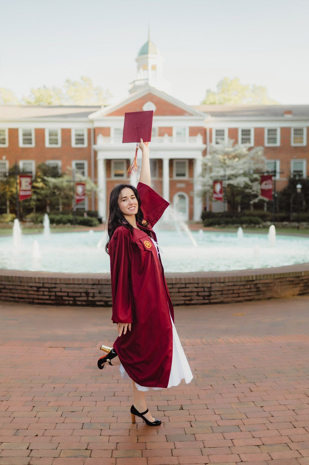 Graduation Photos at Elon University: Celebrating Amelia's Big Moment-Graduation Photos at Elon University Amelia stands in front of Fonville Fountain at Elon University wearing a maroon graduation gown over a white dress, holding her cap and looking thoughtfully to the side.