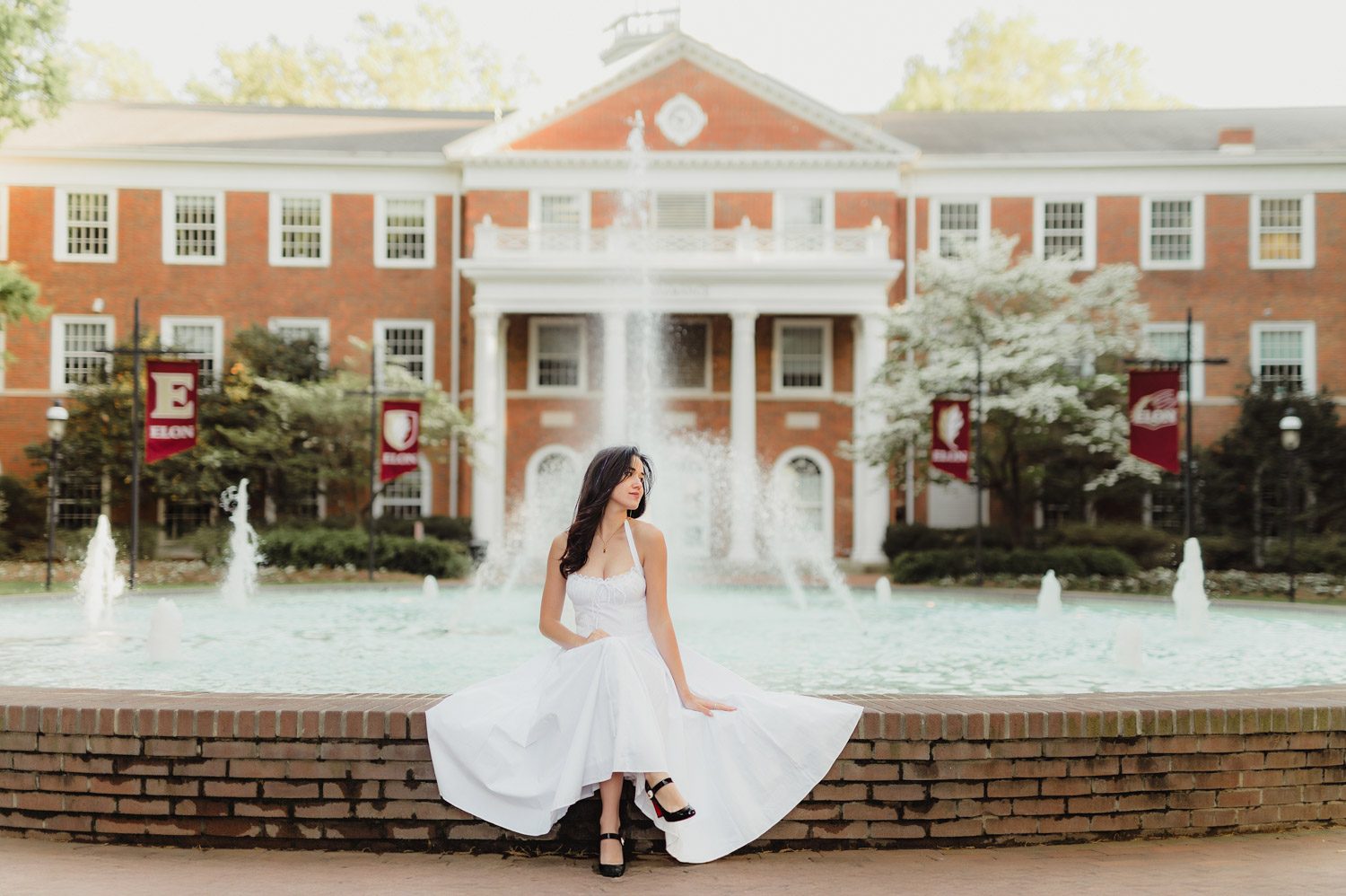 Graduation Photos at Elon University: Celebrating Amelia's Big Moment-Graduation Photos at Elon University Amelia sits gracefully in a white dress in front of Fonville Fountain at Elon University, with Alamance Hall in the background and water jets creating a dreamy atmosphere for Elon graduation portraits.