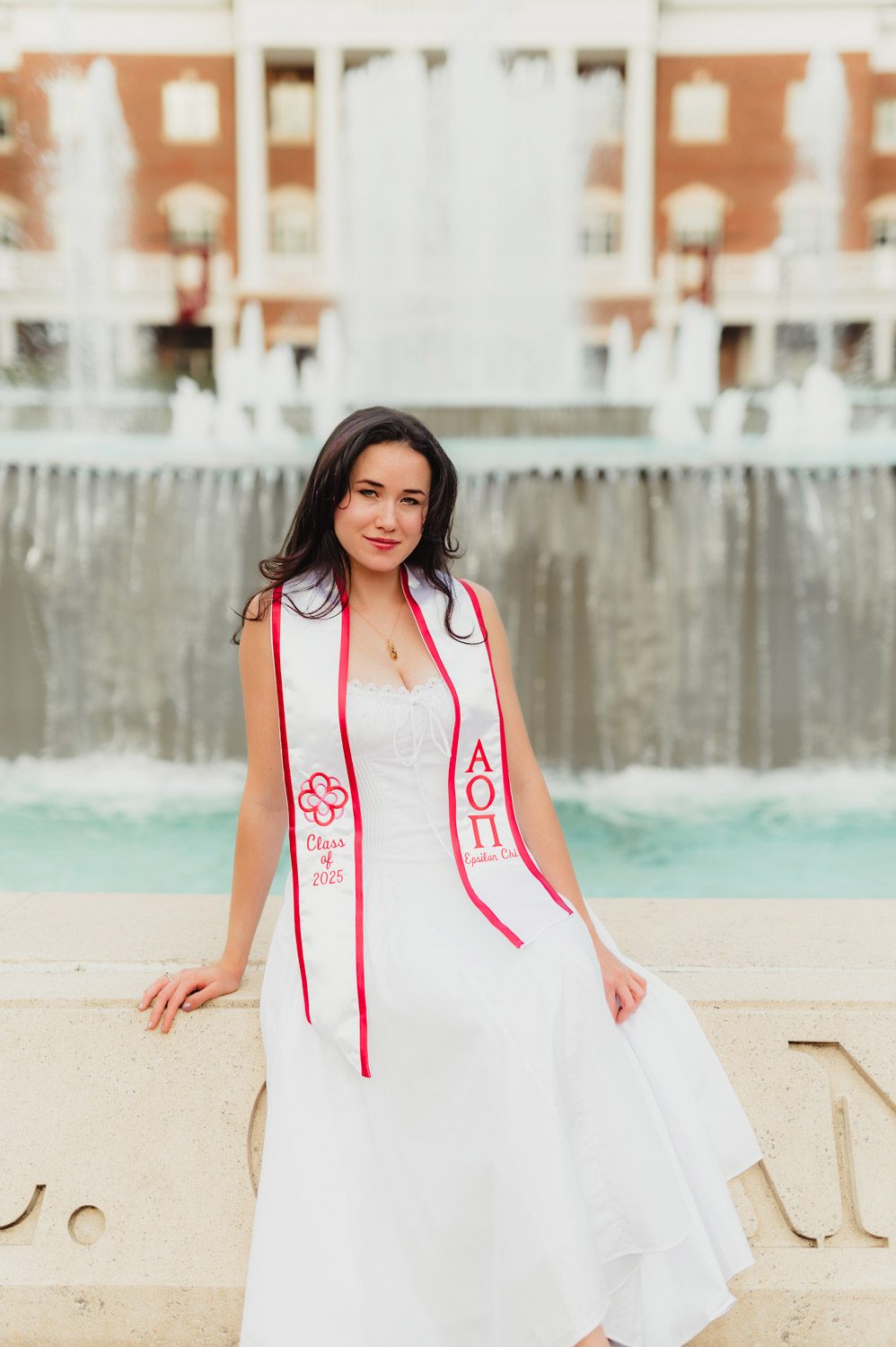 Graduation Photos at Elon University: Celebrating Amelia's Big Moment-Graduation Photos at Elon University Elon University graduate Amelia poses in a white dress and sorority stole in front of the Chandler Fountain, seated on the stone edge with water cascading in the background.