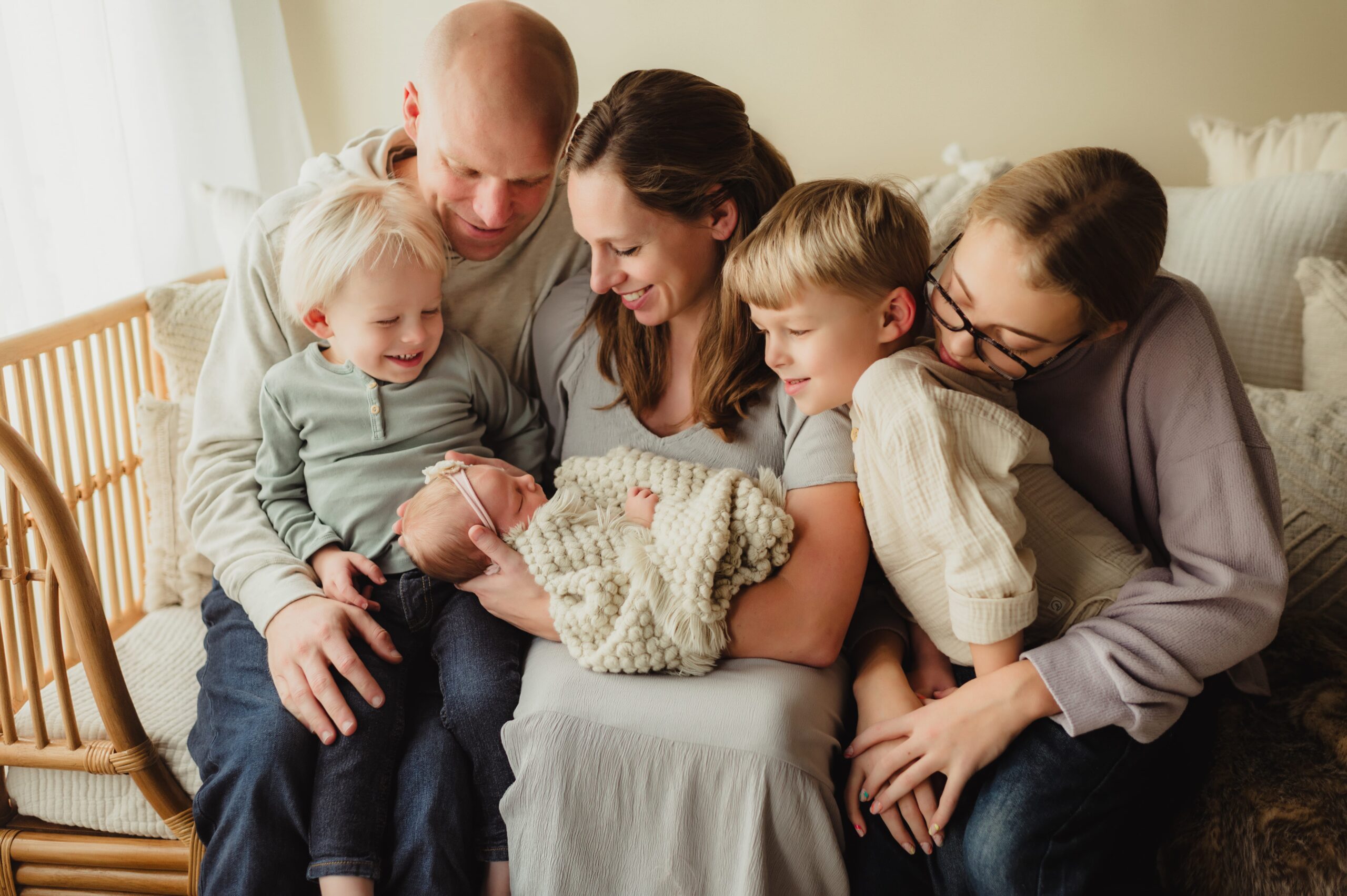 Parents and three siblings admire sleeping newborn baby while sitting on a rattan bed. 