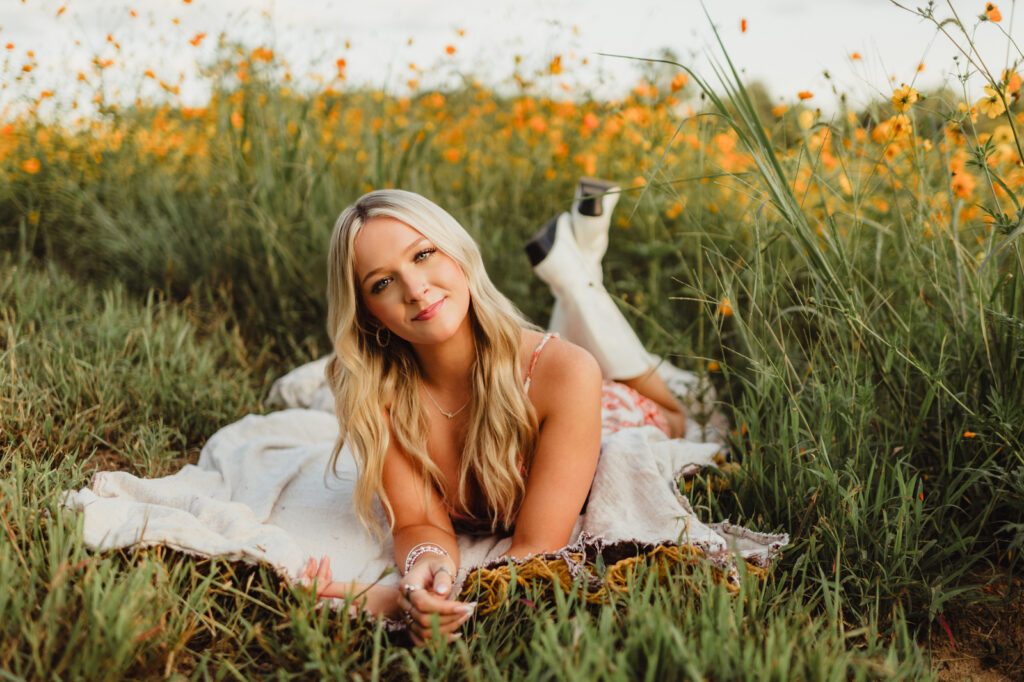 Senior girl lying on a blanket in a wildflower field, propped up on her elbows with a relaxed smile, wearing a patterned sundress and white cowboy boots.