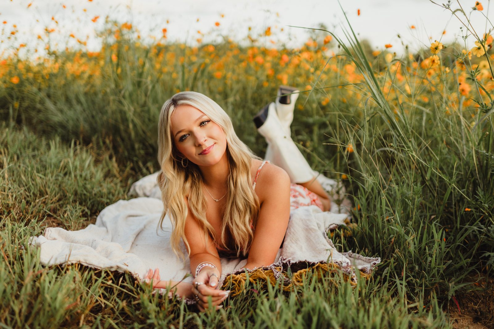 Senior girl lying on a blanket in a wildflower field, propped up on her elbows with a relaxed smile, wearing a patterned sundress and white cowboy boots.