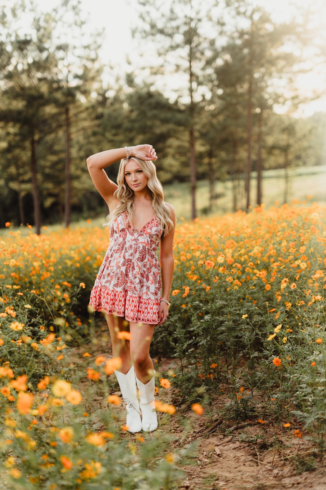 High school senior girl standing in a field of golden wildflowers at sunset, wearing a patterned red sundress and white cowboy boots, posing confidently with one arm lifted above her head.