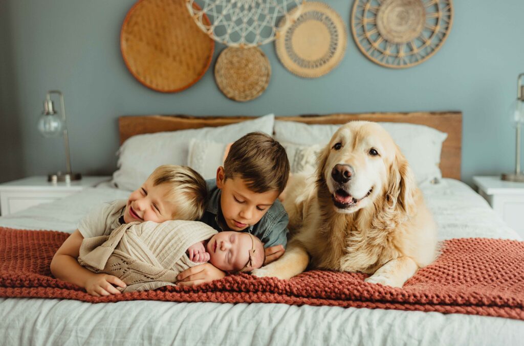 Two brothers lying on the bed with their newborn sibling and golden retriever during an in-home newborn photo session in Greensboro, NC.