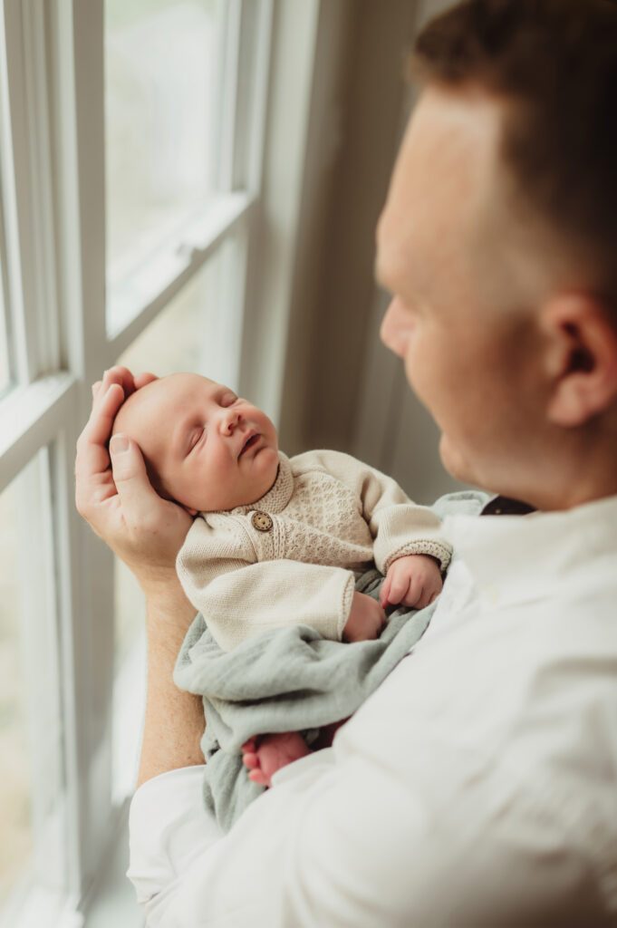 Father holding newborn baby by a window during a natural light in-home newborn photography session in Greensboro, NC.