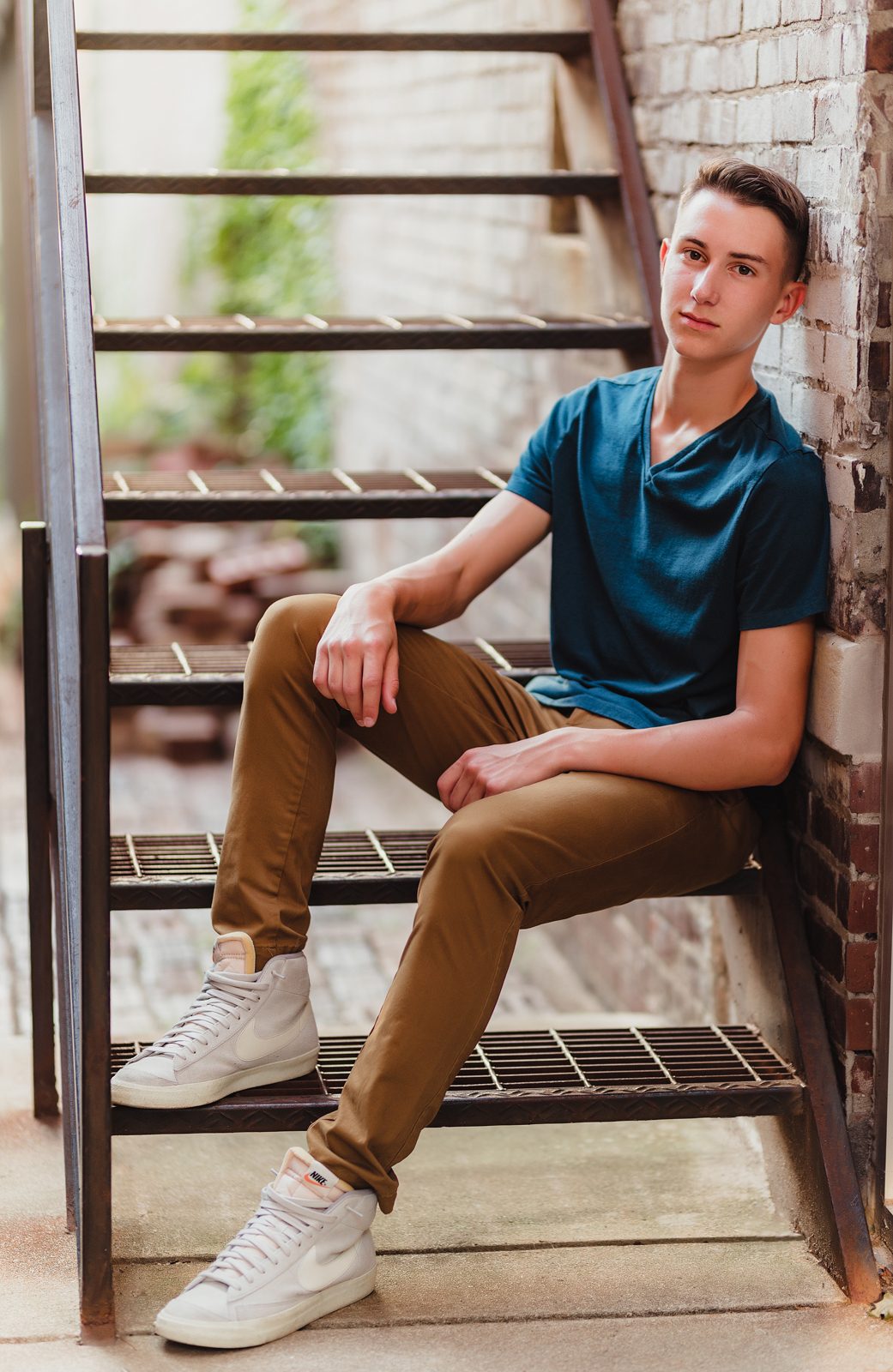 Greensboro Senior Photos That Tell a Story: How to Personalize Your Session-Greensboro senior photographer Senior guy sitting on metal stairs in an alleyway, wearing a teal shirt and brown pants during his urban senior photo session