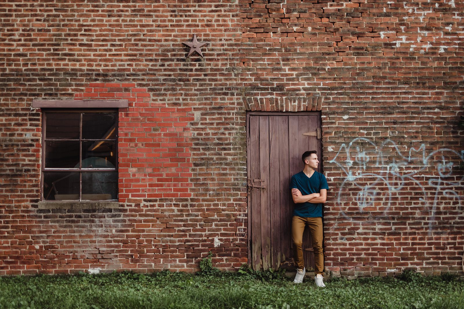 Greensboro Senior Photos That Tell a Story: How to Personalize Your Session-Greensboro senior photographer Senior guy standing against a rustic brick wall with graffiti, wearing a teal shirt and brown pants during an urban senior photo session
