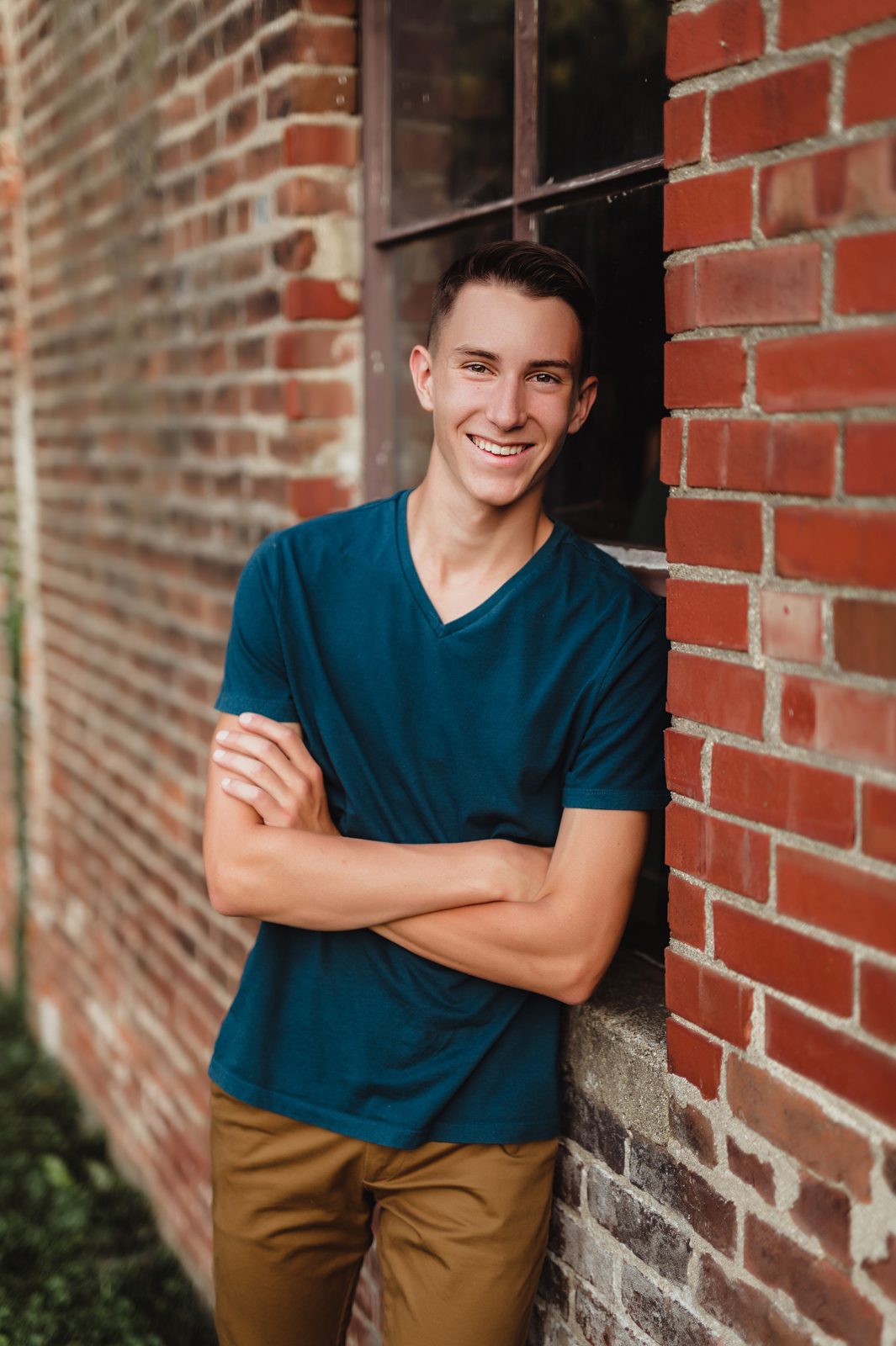 Greensboro Senior Photos That Tell a Story: How to Personalize Your Session-Greensboro senior photographer Senior guy smiling with arms crossed, leaning against a brick wall near a window during his personalized urban photo session