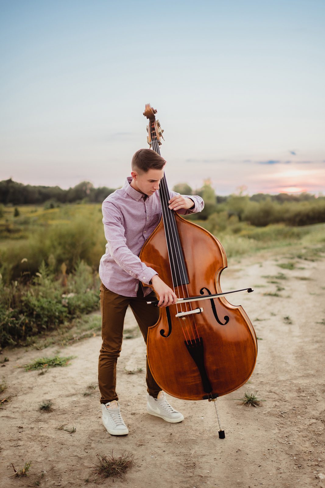 Greensboro Senior Photos That Tell a Story: How to Personalize Your Session-Greensboro senior photographer Senior guy playing upright bass outdoors at sunset, wearing a lavender button-down and brown pants during a storytelling senior session