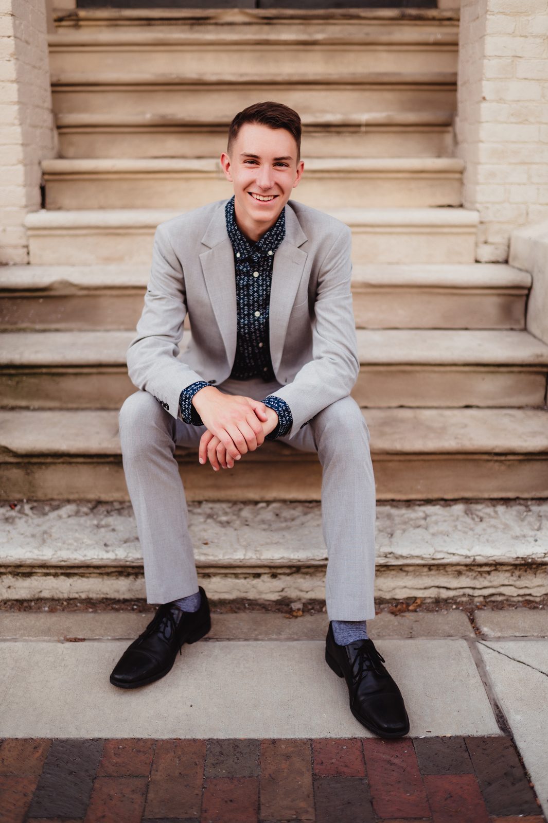 Greensboro Senior Photos That Tell a Story: How to Personalize Your Session-Greensboro senior photographer Senior guy sitting on stone steps, wearing a light gray suit and patterned shirt, smiling during his formal senior portrait session