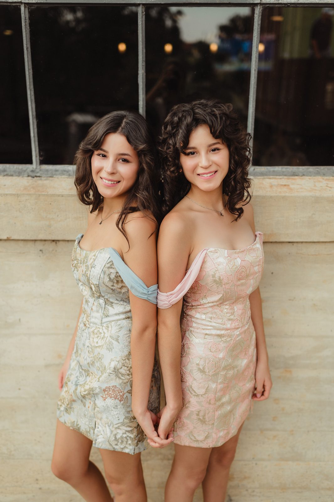 Close-up of twin seniors smiling and holding hands in textured off-the-shoulder mini dresses during fall senior session in Greensboro NC