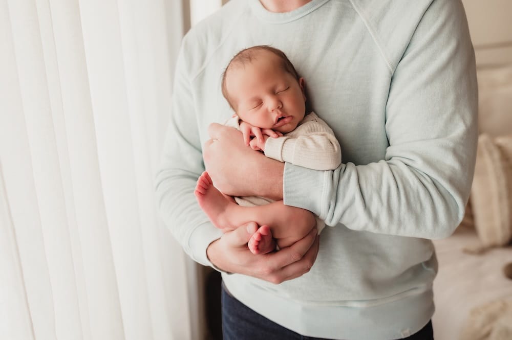 Father gently holding his newborn baby in front of a window during a natural light in-home newborn photo session in Greensboro, NC.