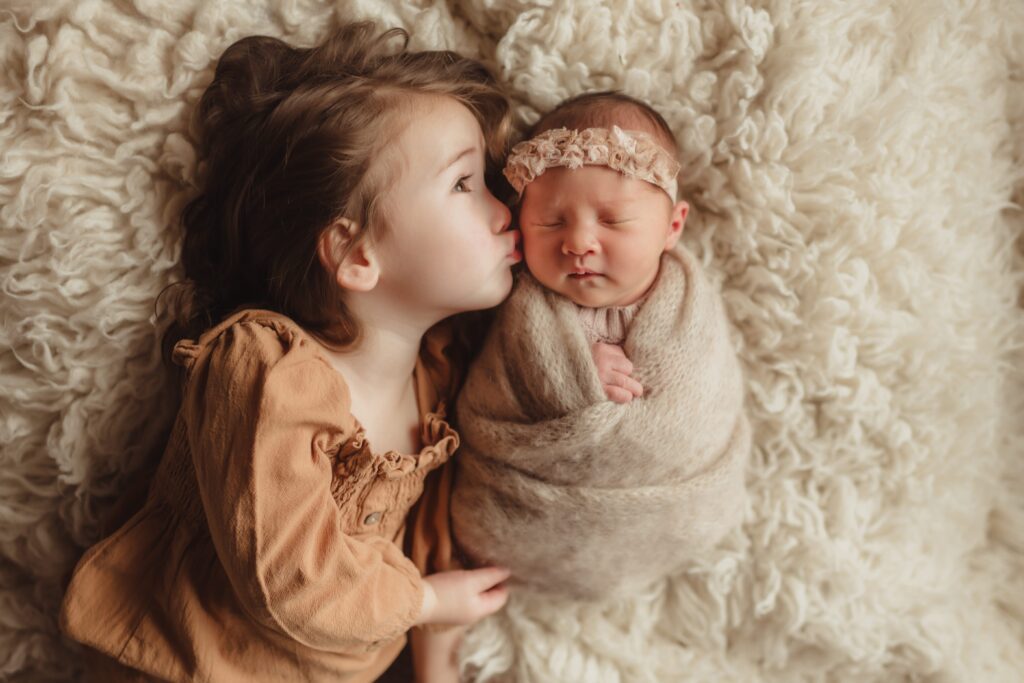 Big sister kissing her newborn baby sibling wrapped in a swaddle during a lifestyle newborn photography session in Greensboro, NC.