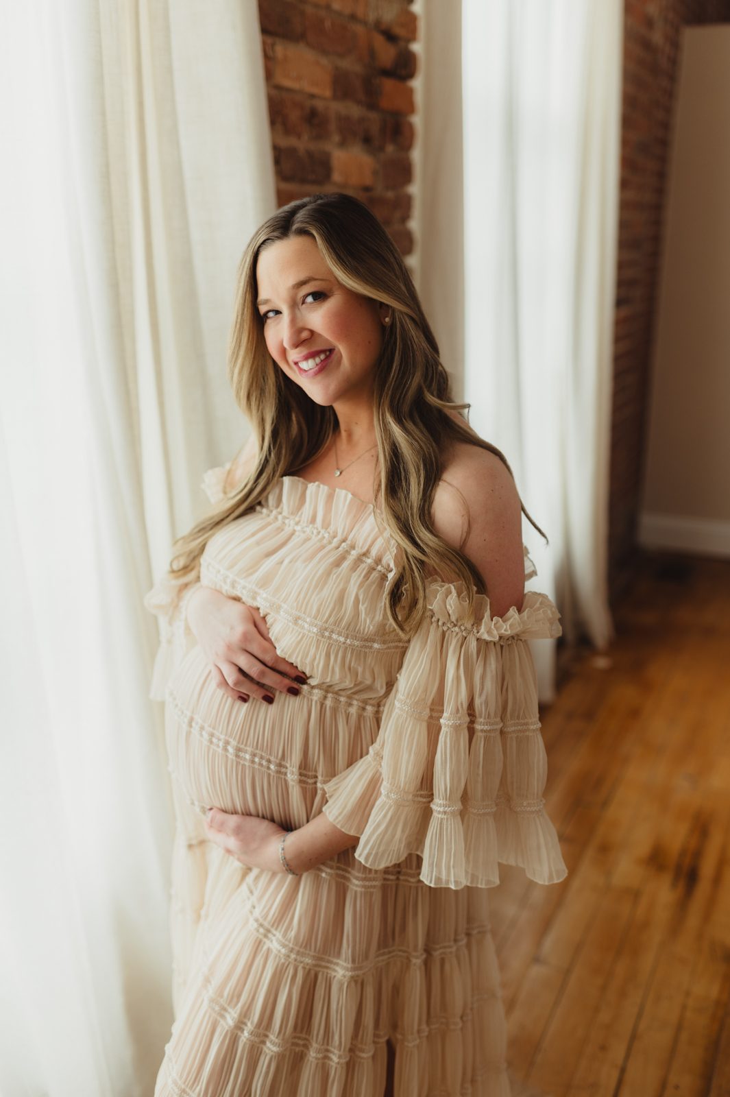 Pregnant mother in cream dress standing by studio window during maternity photos in Greensboro.