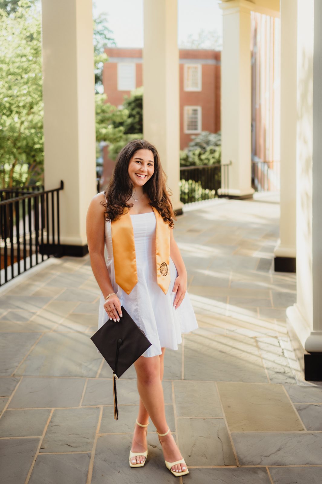 Wake Forest graduation portrait of Mia standing under the colonnade in a white dress and gold stash, holding her cap