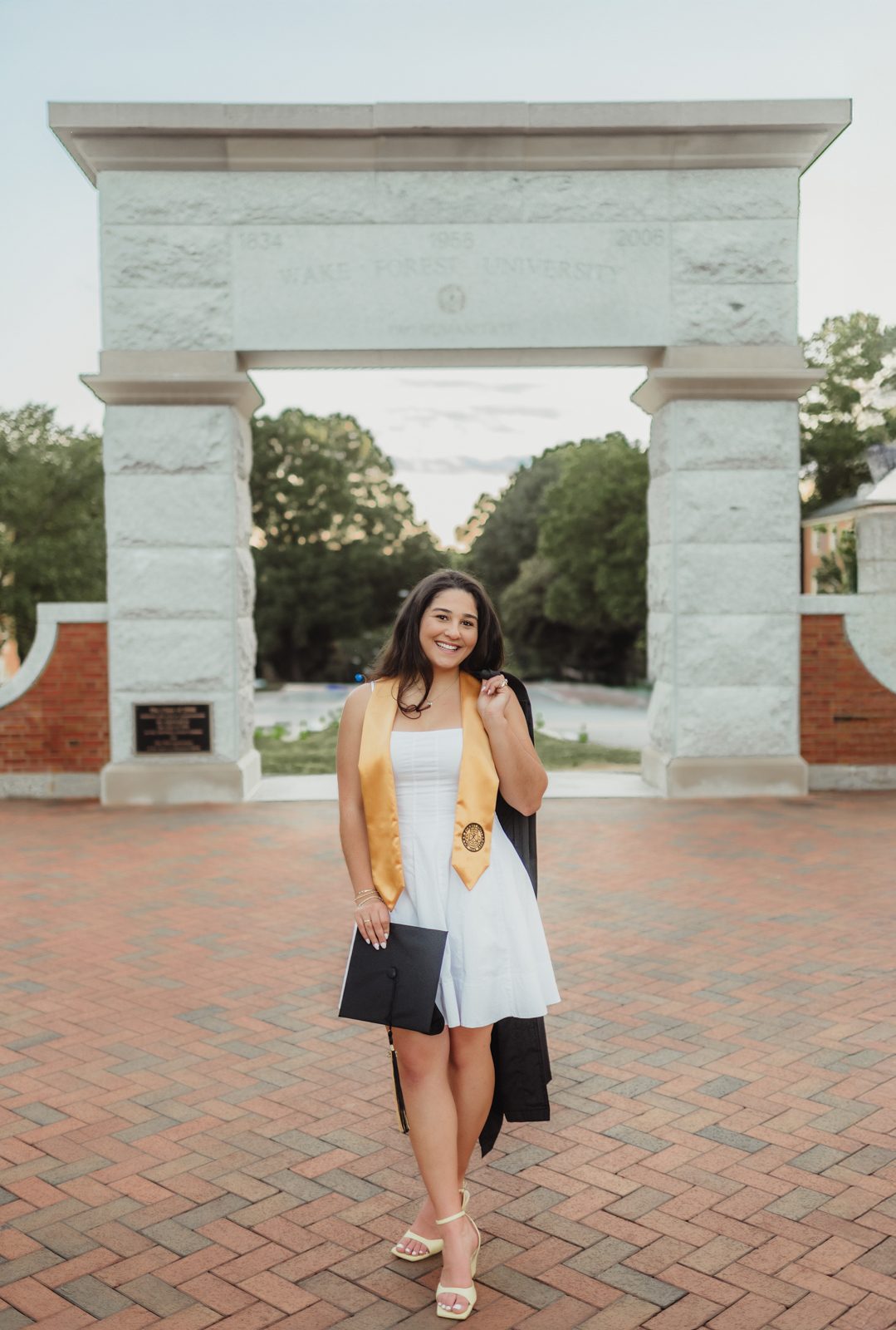 Mia standing confidently in front of the Wake Forest University Stone Arch, holding her cap and gown over one shoulder